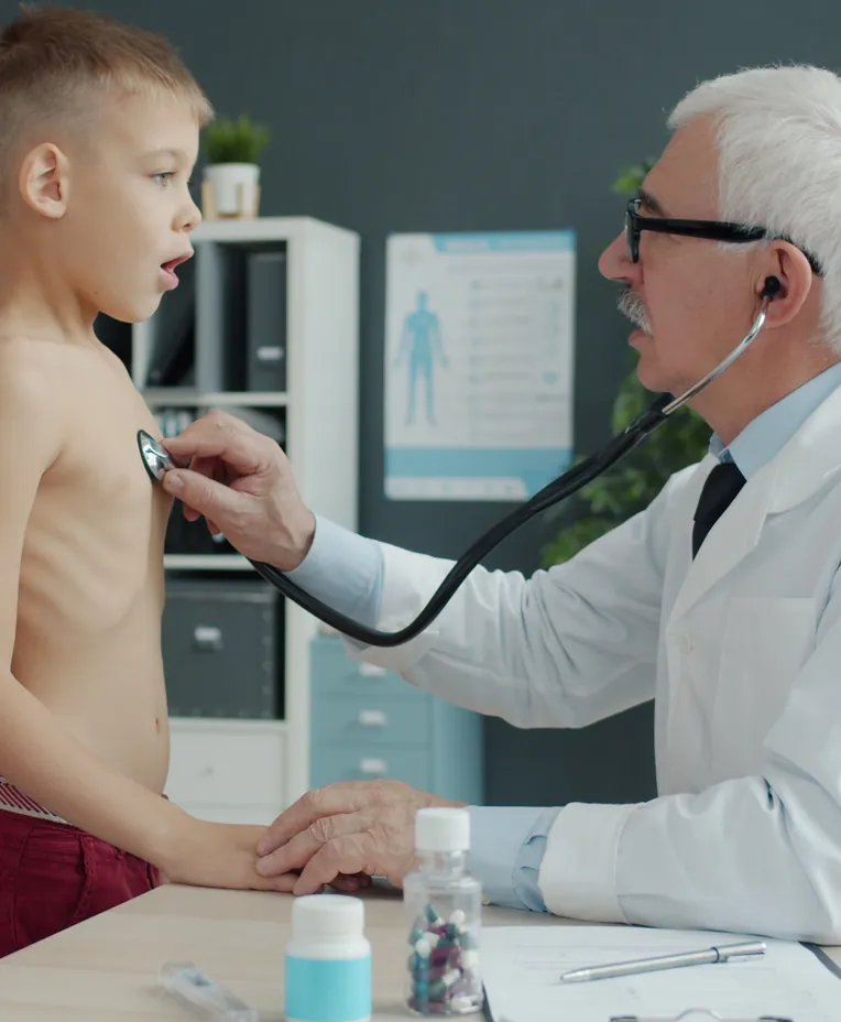 An older doctor listening to a young boy's chest with a stethoscope while his mother looks on