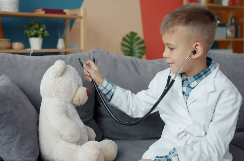 A young boy in a white coat listening to a teddy bear's heart with a stethoscope
