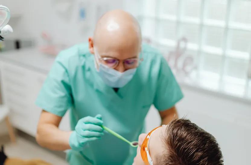 A dentist in green scrubs and a mask examining a young man's teeth in the dental chair