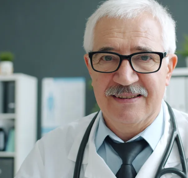 An older doctor with white hair and glasses, in a white coat and tie, smiling in his consulting room