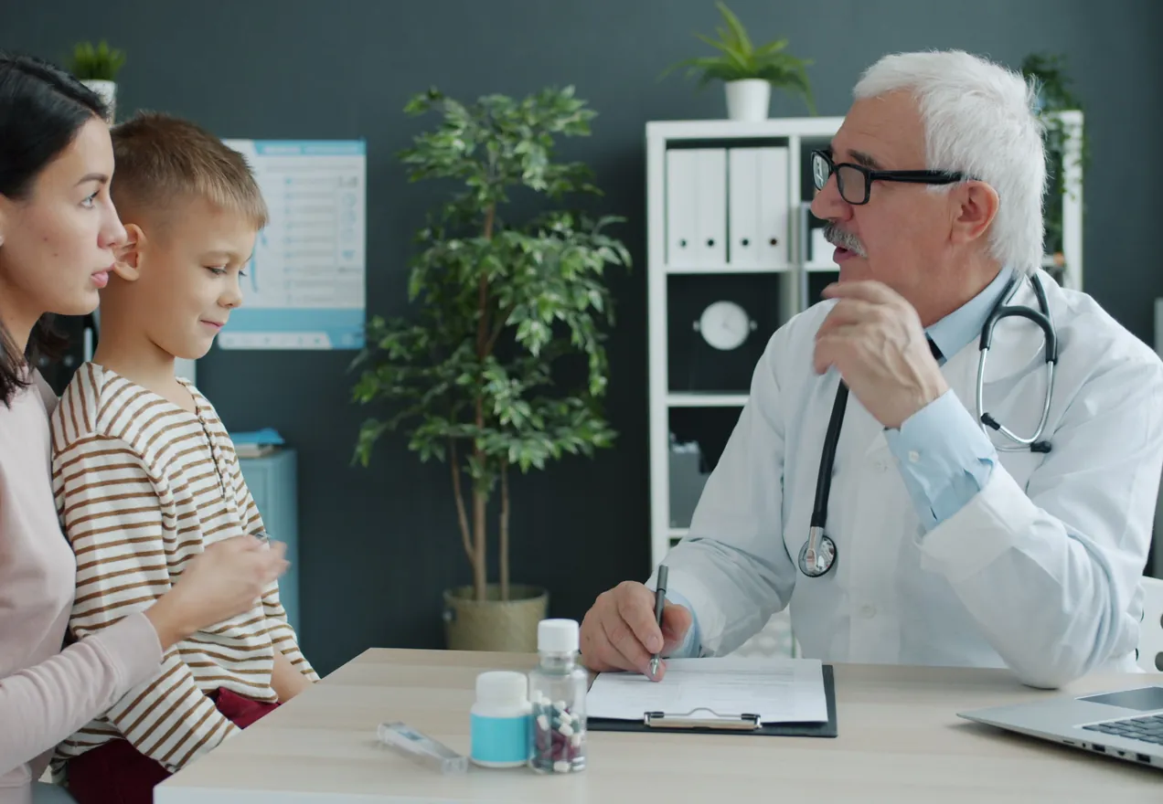 A mother and her young son in a consultation with an older doctor, who is writing notes at his desk