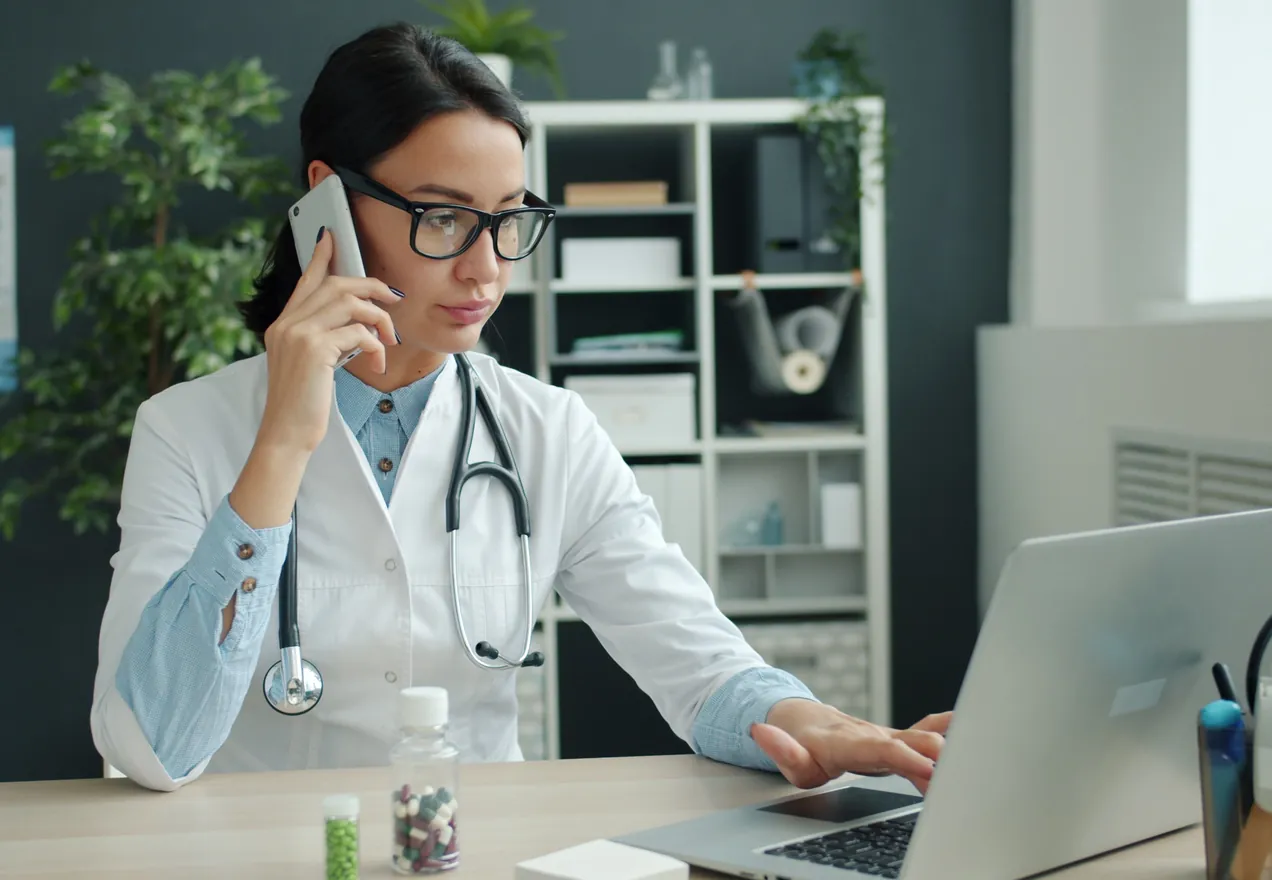 A doctor in a white coat on the phone at her desk, typing on a laptop